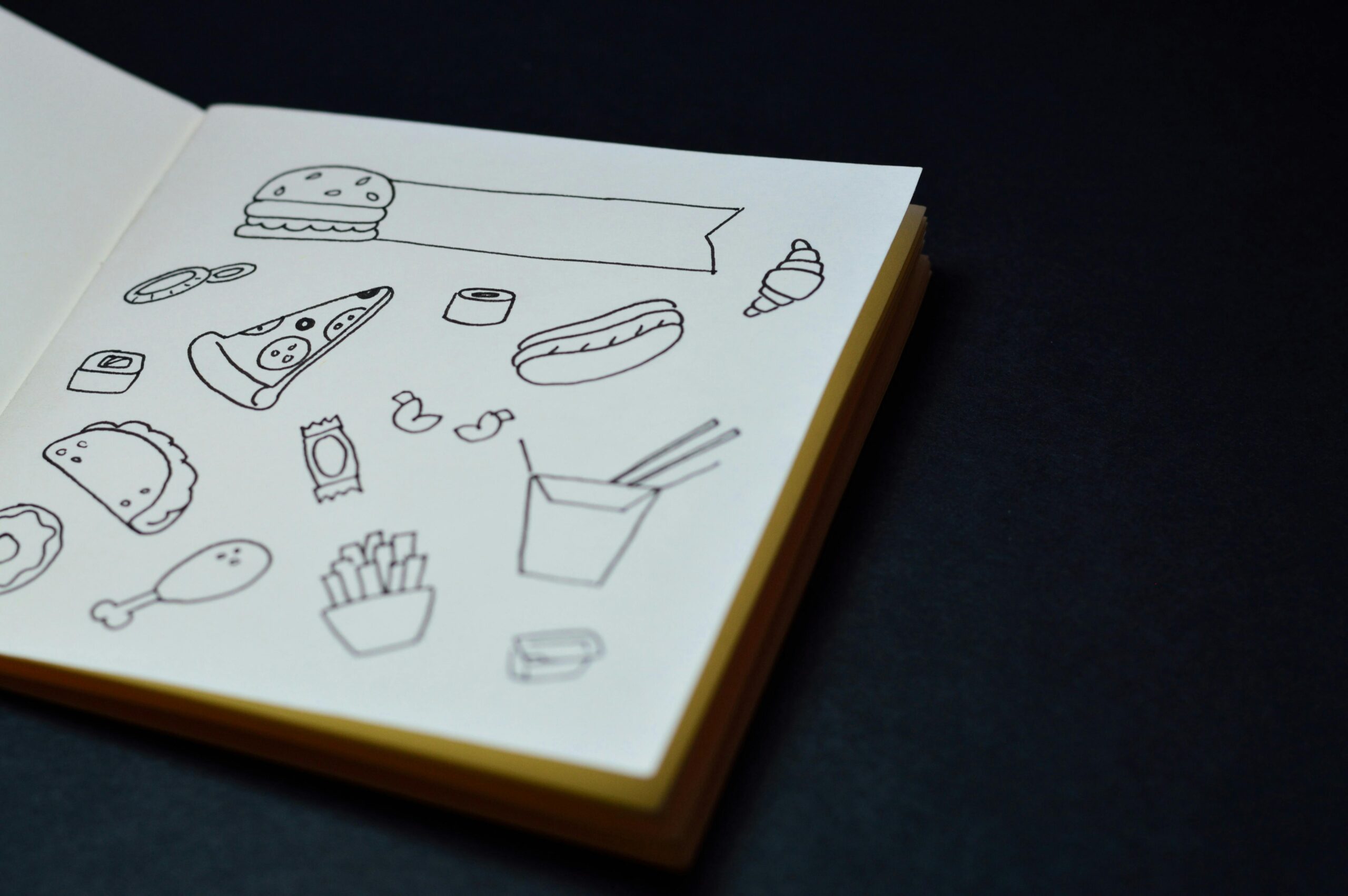 Open sketchbook displaying hand-drawn food doodles like pizza, burger, and fries.art design for project